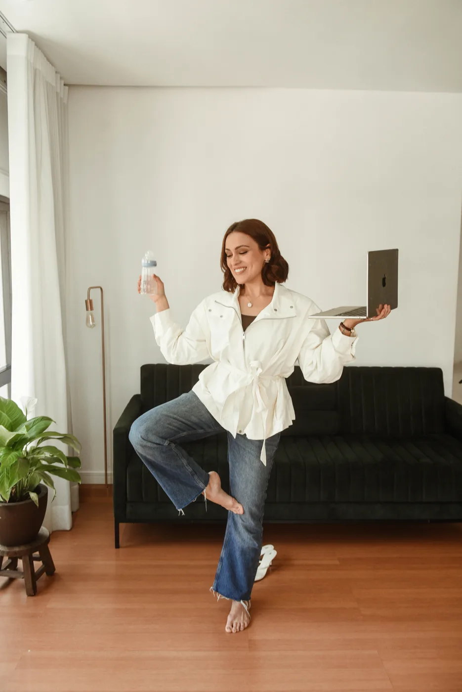 Mulher sorrindo e se equilibrando em um pé só, segurando uma garrafa de água em uma mão e o notebook na outra