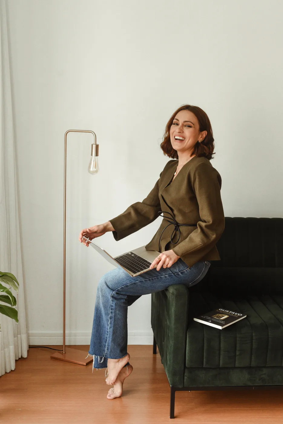 Mulher sorrindo, sentada no braço do sofá com o notebook no colo